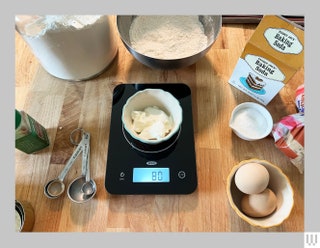 Small black digital scale with a cup on top sitting on a kitchen counter with other ingredients behind