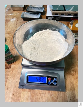 Large silver digital scale with metal bowl of flour on top sitting on a kitchen counter with other ingredients behind