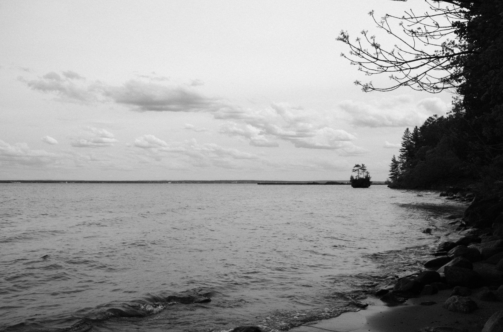 Black and white photo from the edge of a beach with a small island in the distance on a party cloudy day