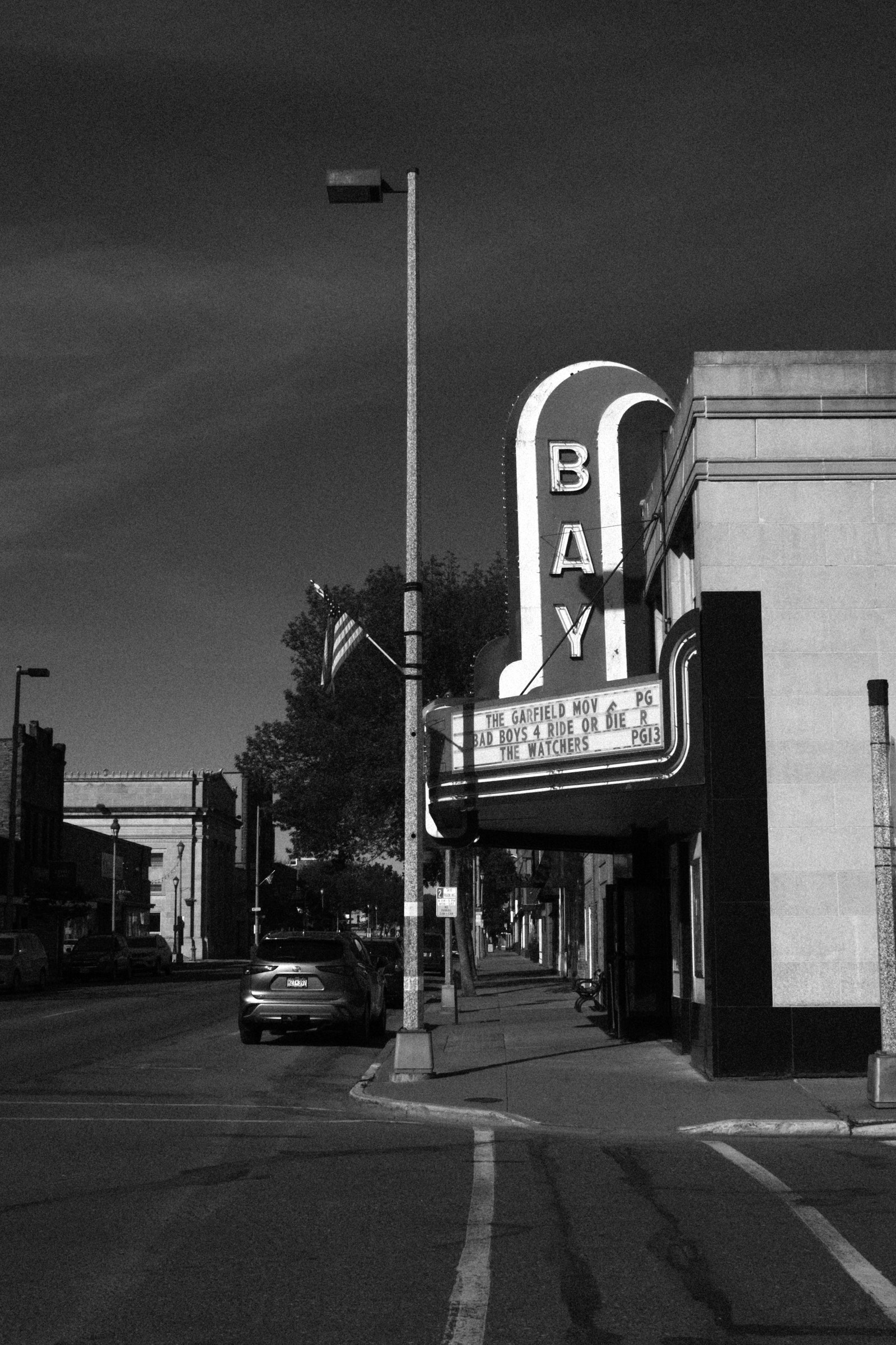 Black and white image of a movie theater sign outside a building with a few cars parked along the sidewalk