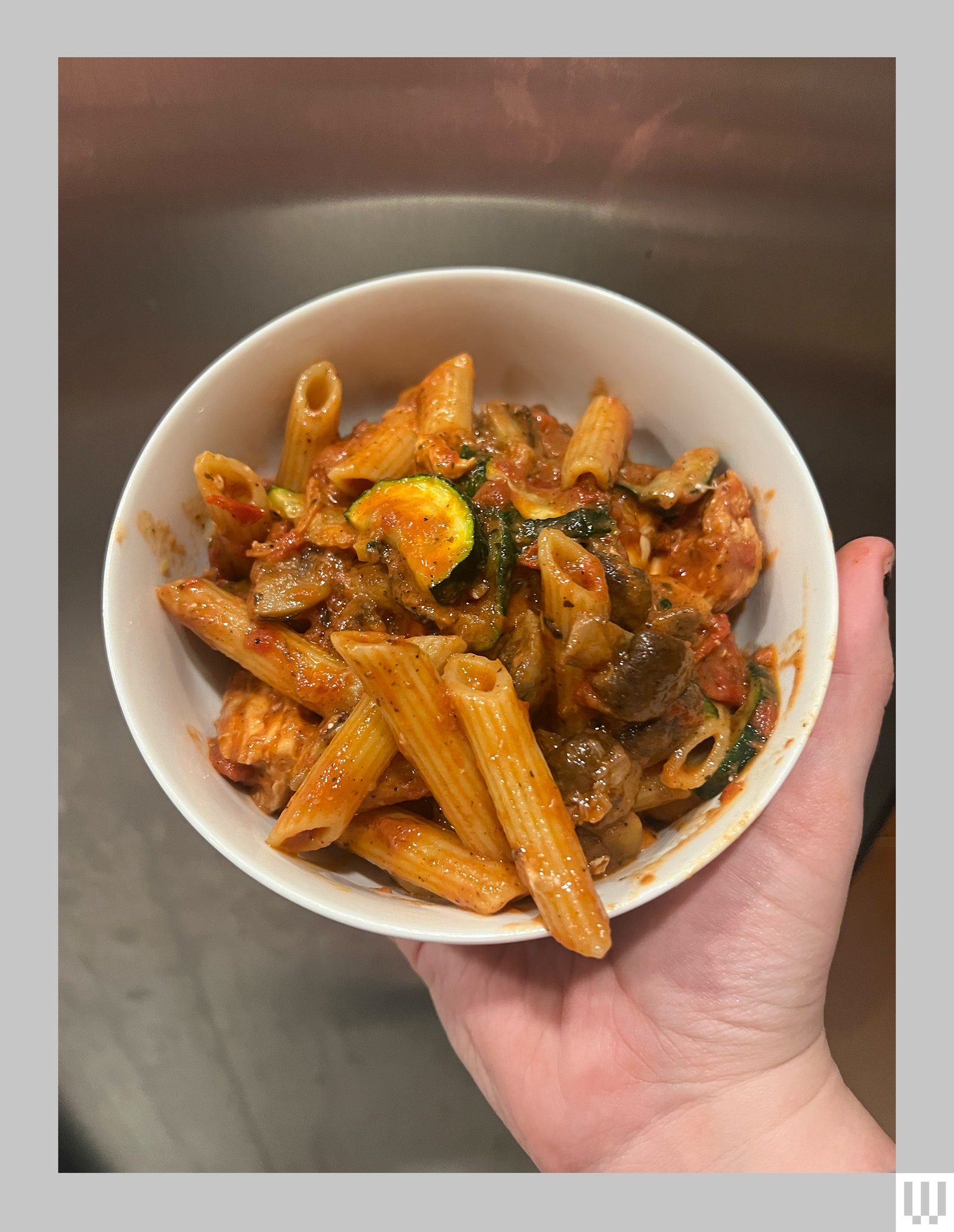 Hand holding a white bowl with pasta and vegetables inside