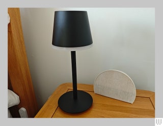 Black lamp beside a semicircle clock both sitting on a wooden bedside table