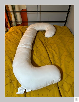 Curvy cshaped white pillow laying across the top of a bed with a yellow blanket underneath