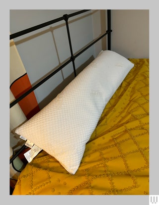 Long white pillow laying across the top of a bed with a yellow blanket underneath