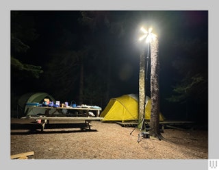 Tall retractable multiprong light illuminating a campsite at night showing off the wooden table supplies and tent