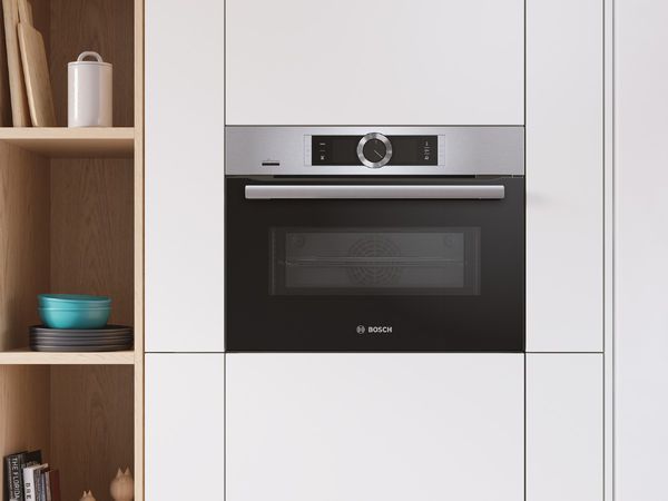 Compact built-in oven on a white kitchen storage wall with open in-wall oak shelves displaying kitchen utensils and cookbooks