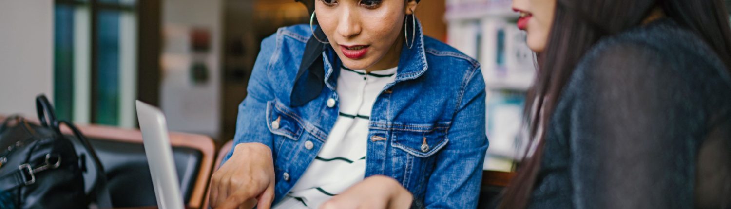 A woman in a jean jacket and black headscarf indicates something on a computer screen to another woman in a black long sleeve shirt.