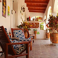 A covered patio with two wooden chairs features patterned cushions, potted plants, and wall decorations. Red ceiling beams and brick flooring complete the cozy atmosphere.