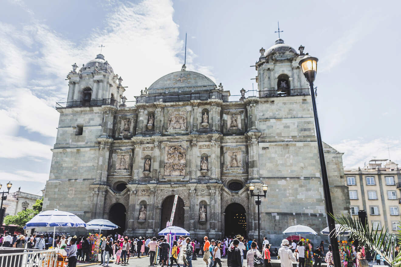 People gather outside a historic stone cathedral on a sunny day, with some using umbrellas for shade.