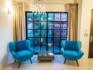 Two turquoise chairs with blue cushions face each other in front of a large window with beige curtains. A small coffee table with books and a vase of flowers sits between the chairs.