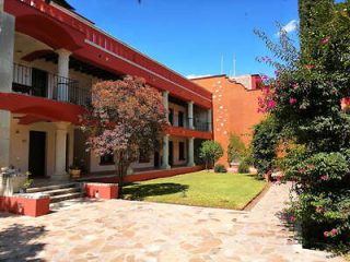 A two-story building with red and white exterior next to a sunny courtyard, featuring paved stones, grass, a small tree, and flowering shrubs.