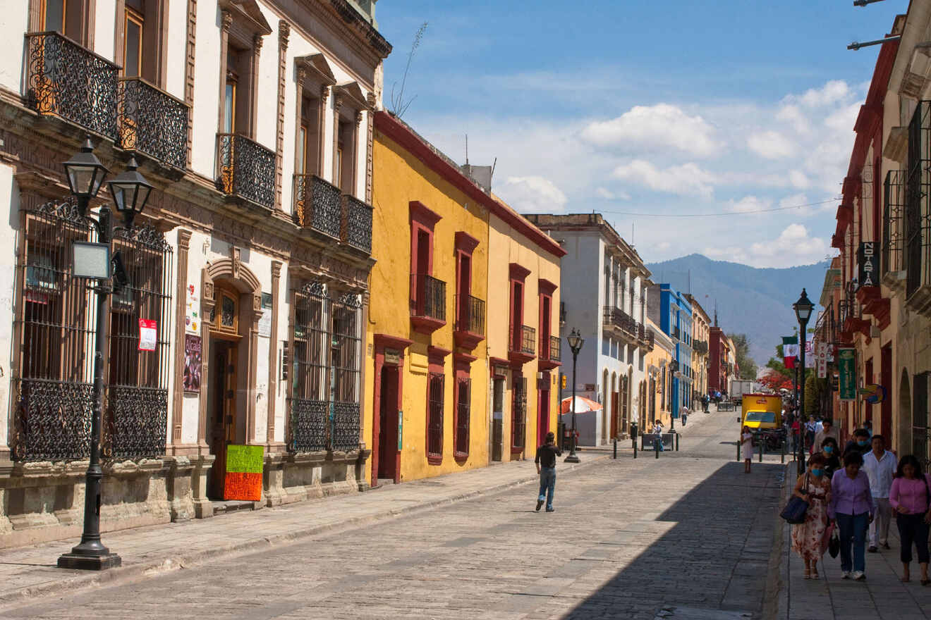 A street scene with colorful buildings and ornate balconies, people walking, and a mountain in the background under a partly cloudy sky.