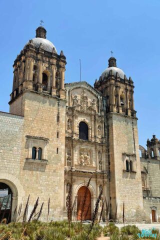 Front view of an old stone church with two dome-topped towers and ornate facade details under a clear blue sky.