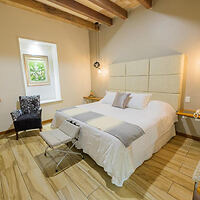 A cozy bedroom with a large bed, wooden ceiling beams, a small window with natural light, and a modern chair.