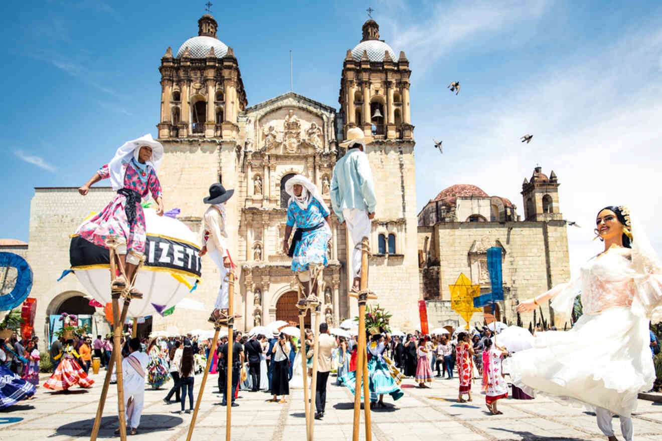 People in traditional attire perform on stilts in front of a historic cathedral. A festive crowd and pigeons are in the background.