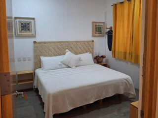 A small bedroom with a queen bed, white bedding, and yellow curtains. Two framed artworks are on the wall. A side table with a plant is next to the bed.