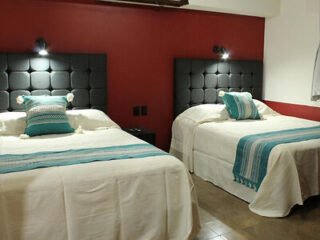 Two neatly made beds with patterned pillows and throws, set against a red wall with overhead lighting.