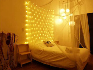 A cozy bedroom with warm string lights on the wall, a bed with white linens, and hanging pendant lights. A side table holds decorative vases with branches. A net canopy partially encloses the bed.