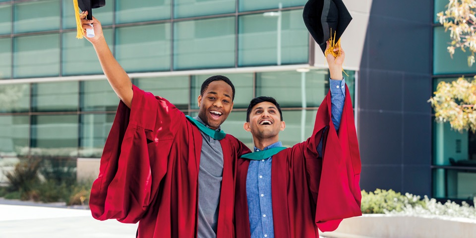 Two students in their graduation robes smiling while holding their hats in their hats in celebration of graduating.