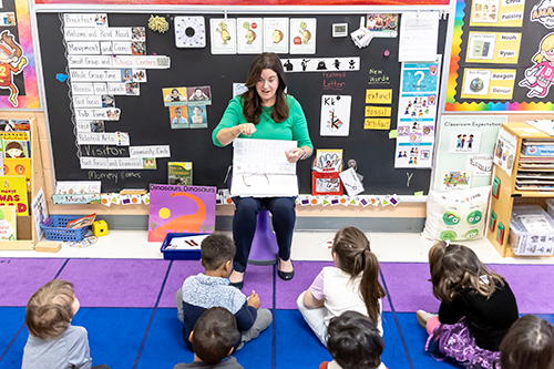 a teacher at Bushy Park ES sits in a chair while pre-k students sit on the floor in front of her