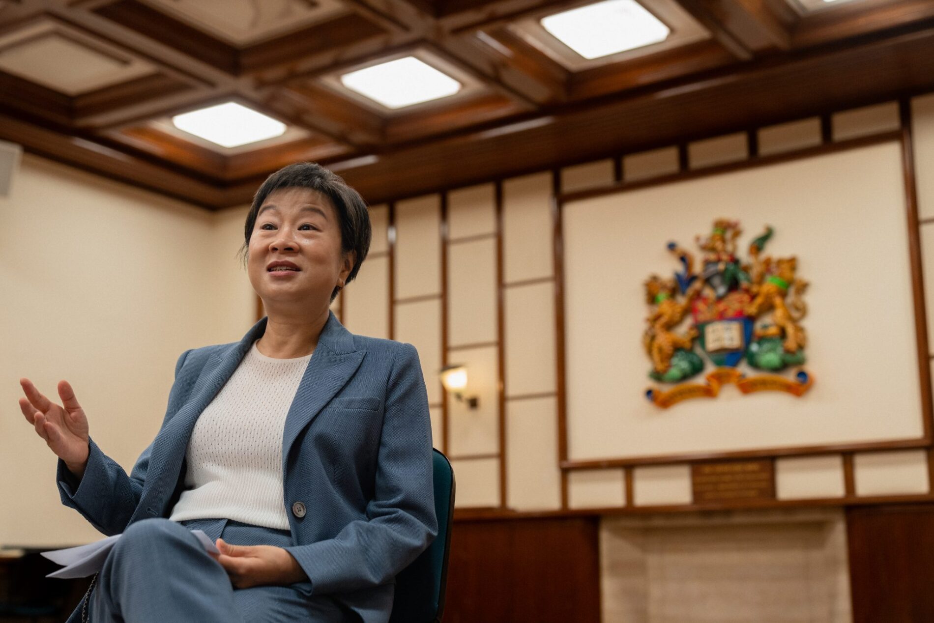 Photo of a lady in animated dialogue sitting in front of a wall-mounted crest of The University of Hong Kong