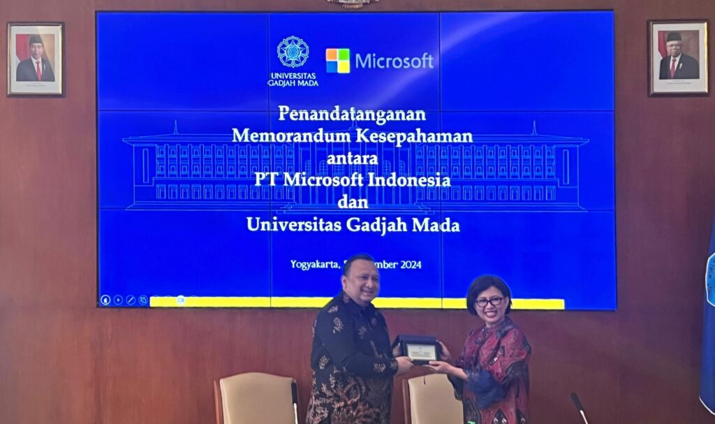 Two individuals stand in front of a large screen displaying the logos of Gadjah Mada University and Microsoft, indicating a signing of a memorandum of understanding between Microsoft Indonesia and Gadjah Mada University.