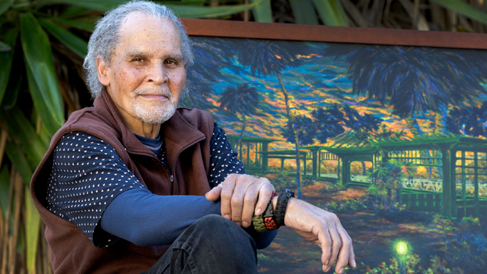 George Collins sits with his hands draped over his knees in front of one of his paintings