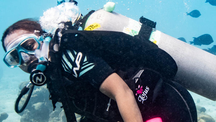 A woman swims underwater while wearing an oxygen tank, breathing mask and wetsuit.