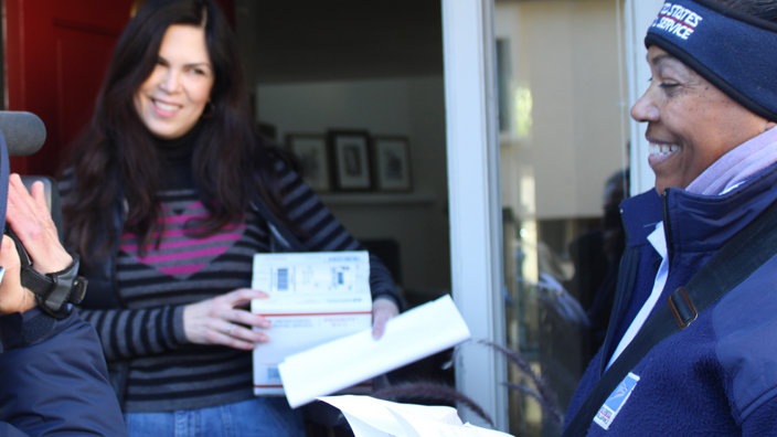 A woman receives a package at her front door from a USPS letter carrier.