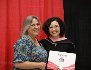 A graduate with brown hair and glasses smiles at the camera while posing with another woman