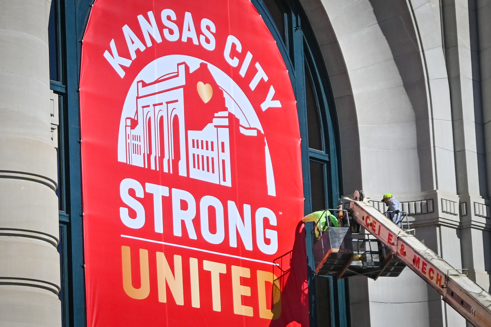 Two men can be seen high off the ground in a bucket lift that is extended toward the outside of a large, concrete building. They appear to be hanging a large red banner with white and gold letters that reads "Kansas City, Strong, United." 