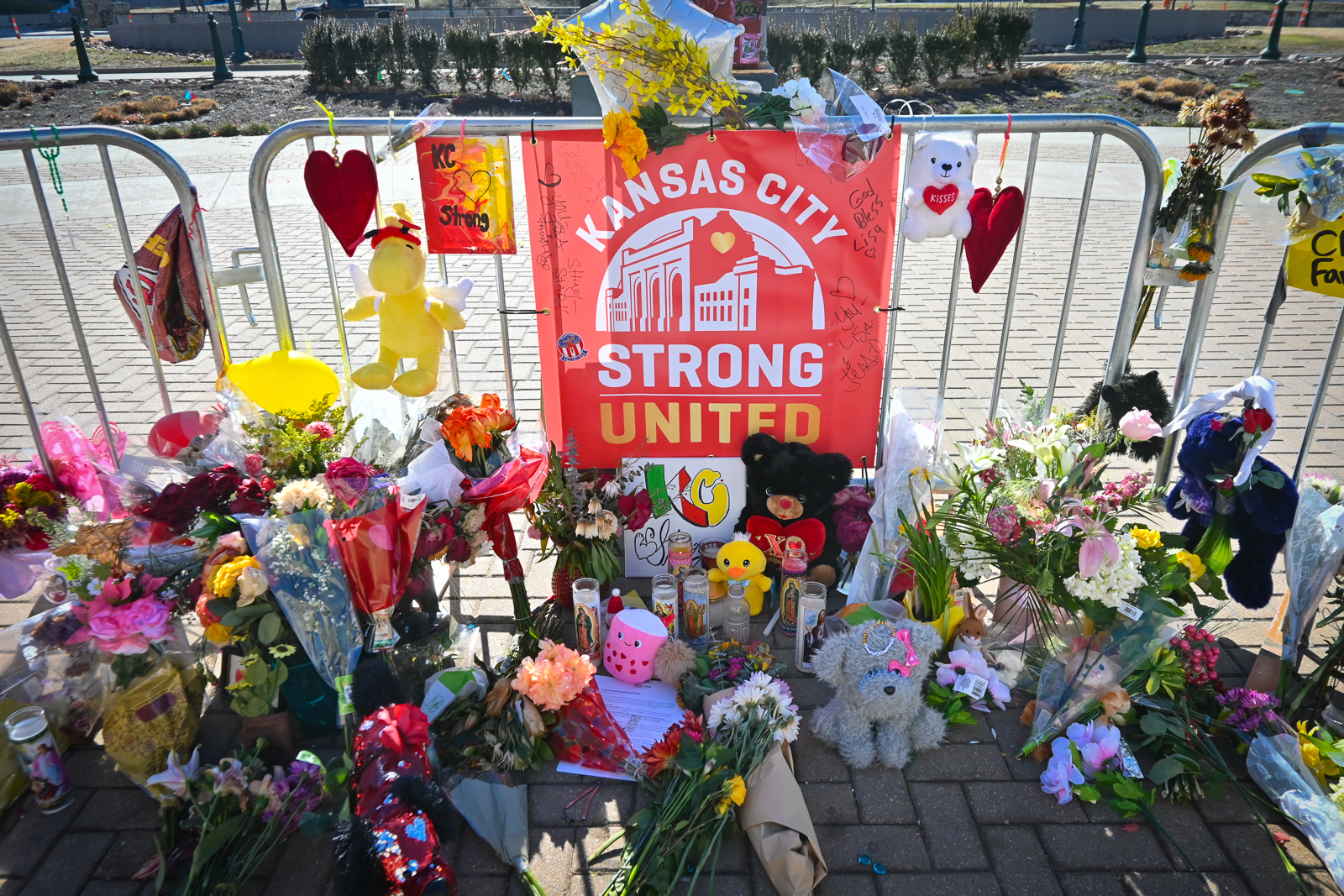 Outdoor photo in bright sunlight of a metal barrier that is lined with red and yellow balloons. There are flowers gathered on the pavement in front of it along with small stuff animals. A sign attached to the metal barrier reads "Kansas city Strong, United."