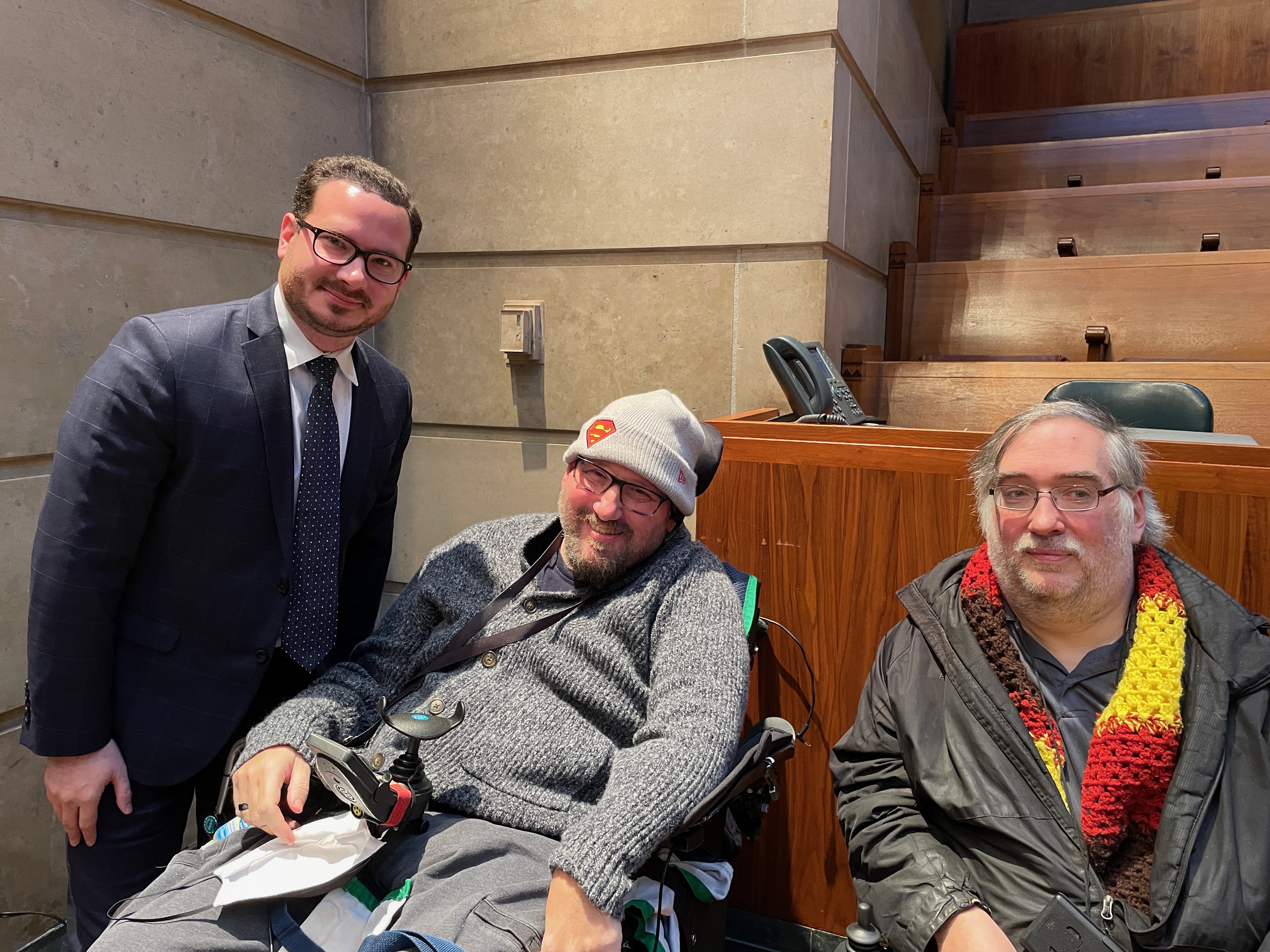 Mitch Nowakowski stands to the left. He is wearing a suit and tie, glasses, and has short brown hair and a short beard. BJ Stasio sits in his power wheelchair in the middle of the photo. He has a beard, and is wearing glasses, a grey sweater, grey pants, and a superman hat. Todd Vaarwerk sits next to BJ in a power wheelchair and has grey-brown hair and a beard. He is wearing glasses, a black jacket, a multicolored scarf, and a black shirt. 