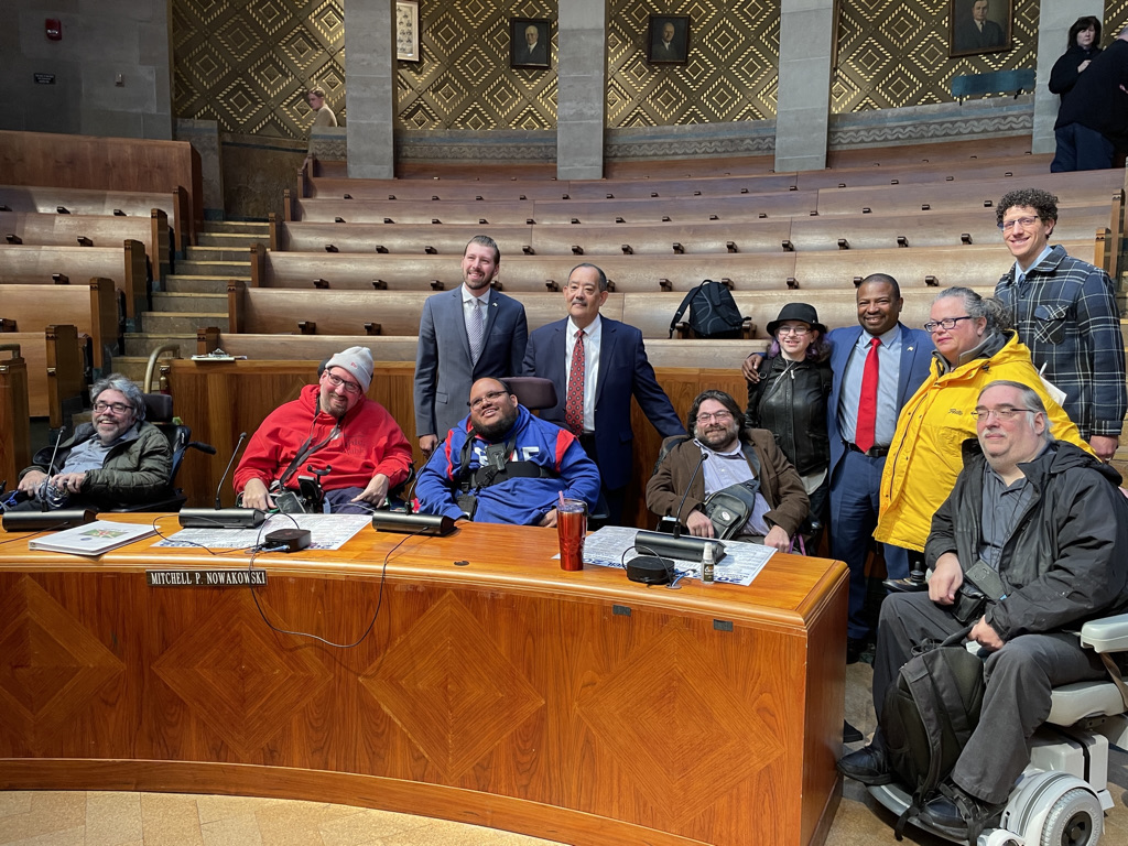 Several people stand or sit in wheelchairs for a photo in a wood and stone government council chamber. 