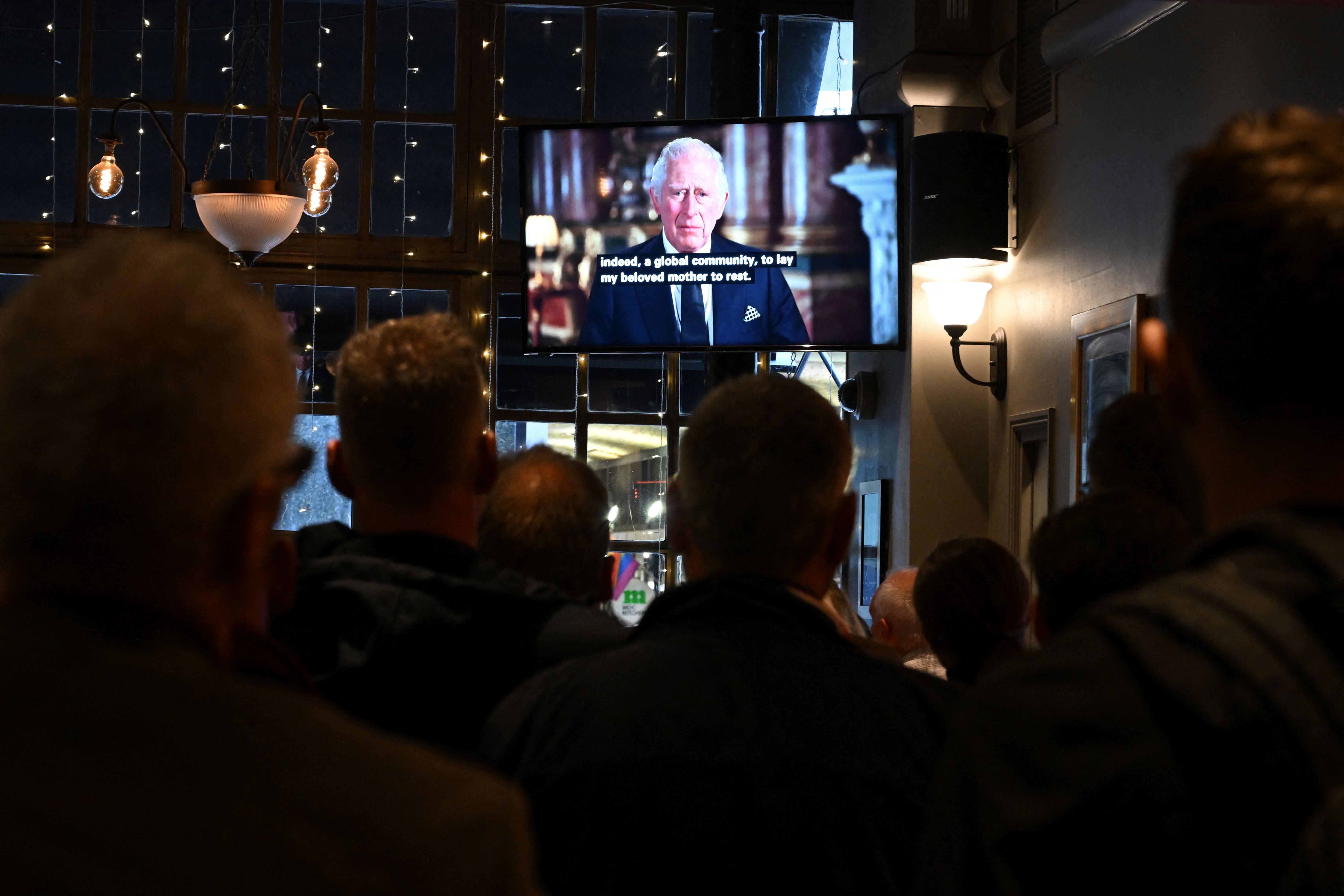 People watch King Charles speak on the television in a dark pub.