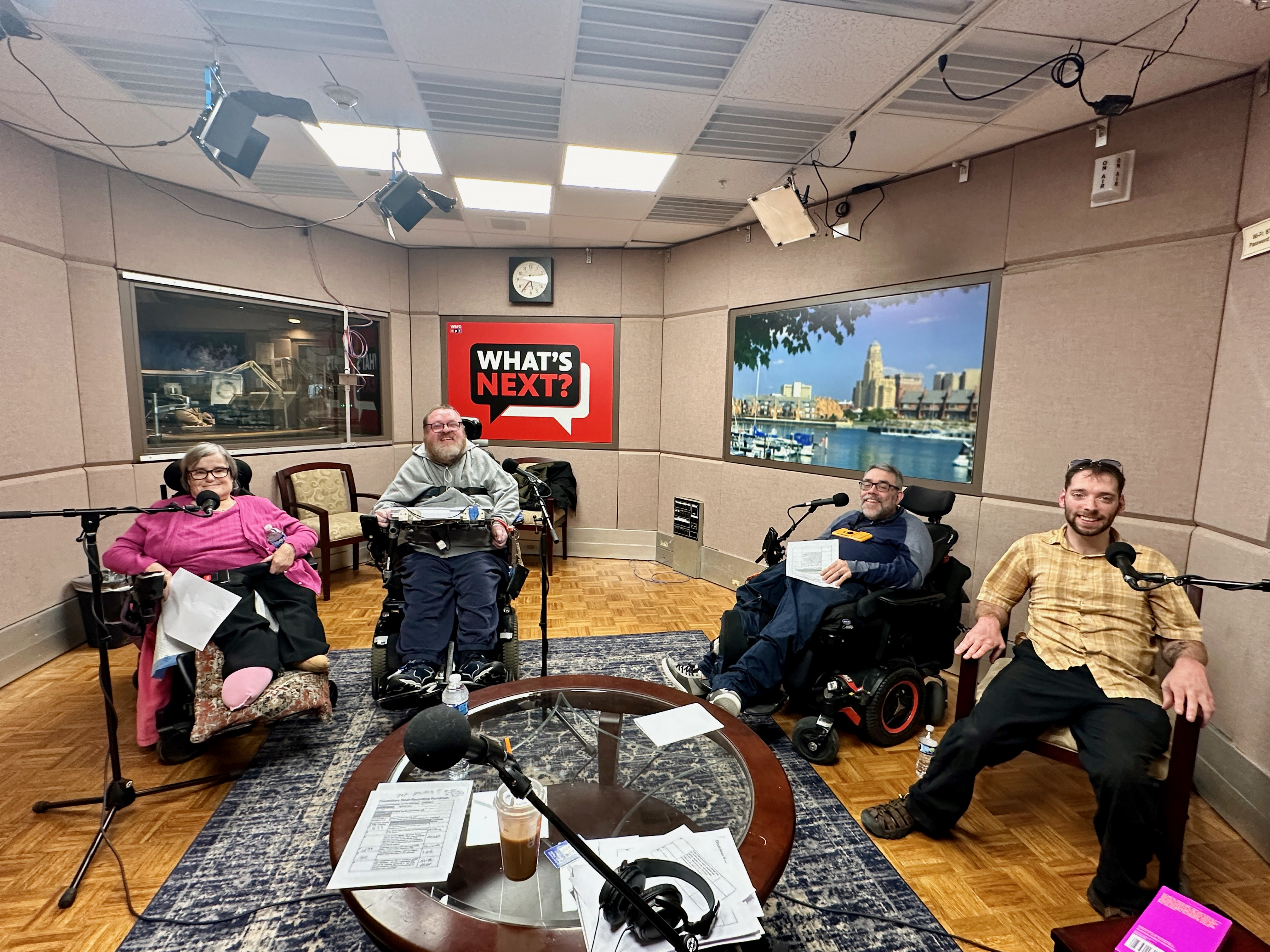 From left to right is Gwen Squire, Lee Allan Hartlieb, Mike Rogers and Alex Jusko sitting in a large studio at WBFO. Behind them you can see into another radio studio, the WBFO What's Next? logo on the wall, and a blurred image of downtown Buffalo. Gwen, Lee and Mike all use power wheelchairs and Alex sits in a studio chair. There is a table and microphones in front of them. Gwen has short brown-grey hair, wears glasses, and is wearing a pink shirt and cardigan with black pants. Lee has dirty blonde hair and a beard, wears glasses, and is wearing a grey sweatshirt, dark pants and sneakers. Lee also has a computer attached to his wheelchair that is used for communication. Mike has dark brown-grey hair and a beard, wears glasses, and is wearing a dark blue and grey jacket with dark blue pants and sneakers. Alex has brown hair and a beard and is wearing a yellow and brown plaid button down short sleeve shirt with black pants. 