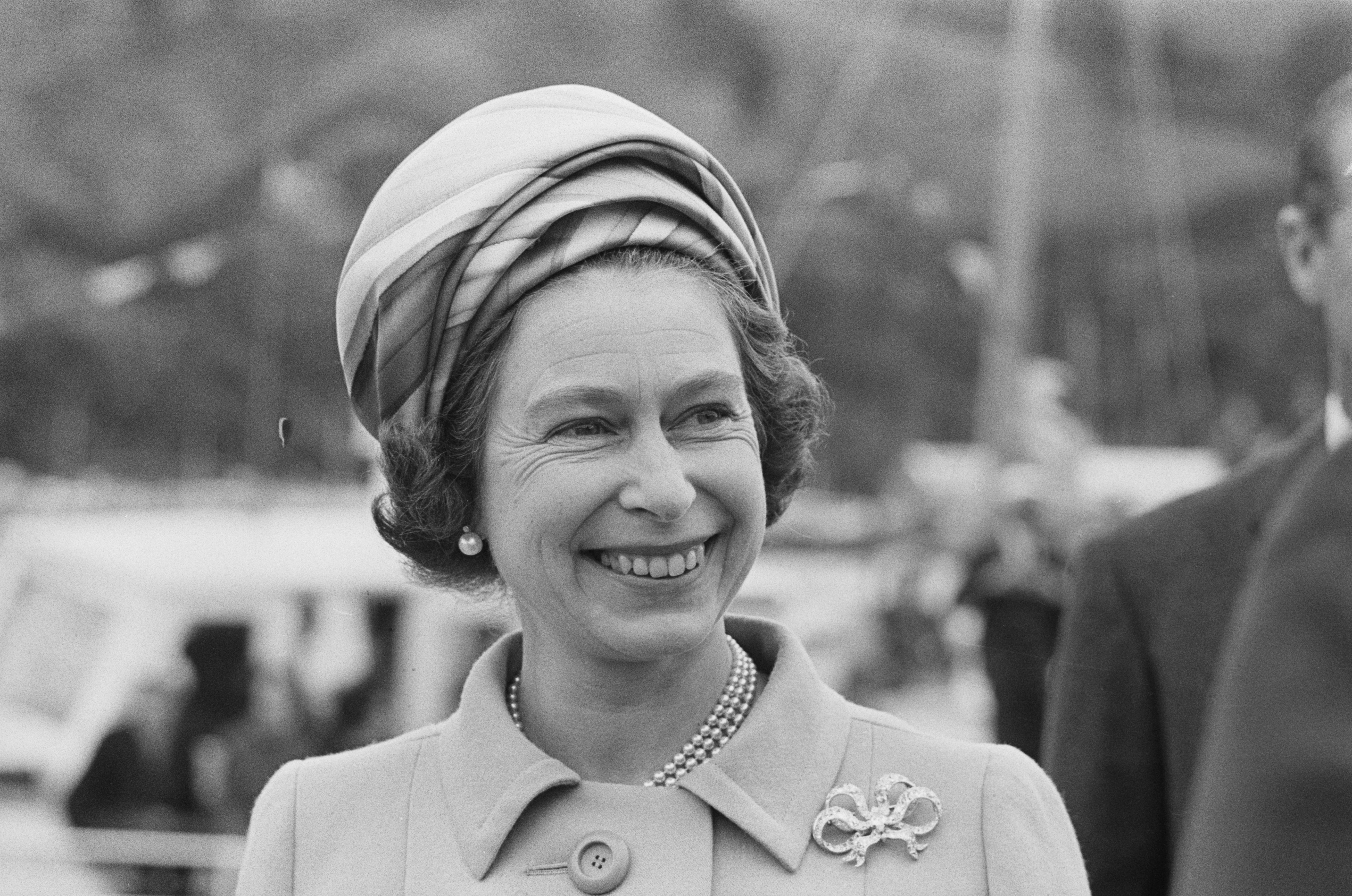 A black and white photo of a similing Queen Elizabeth, wearing a jacket, pearl necklace and head scarf, in 1970.
