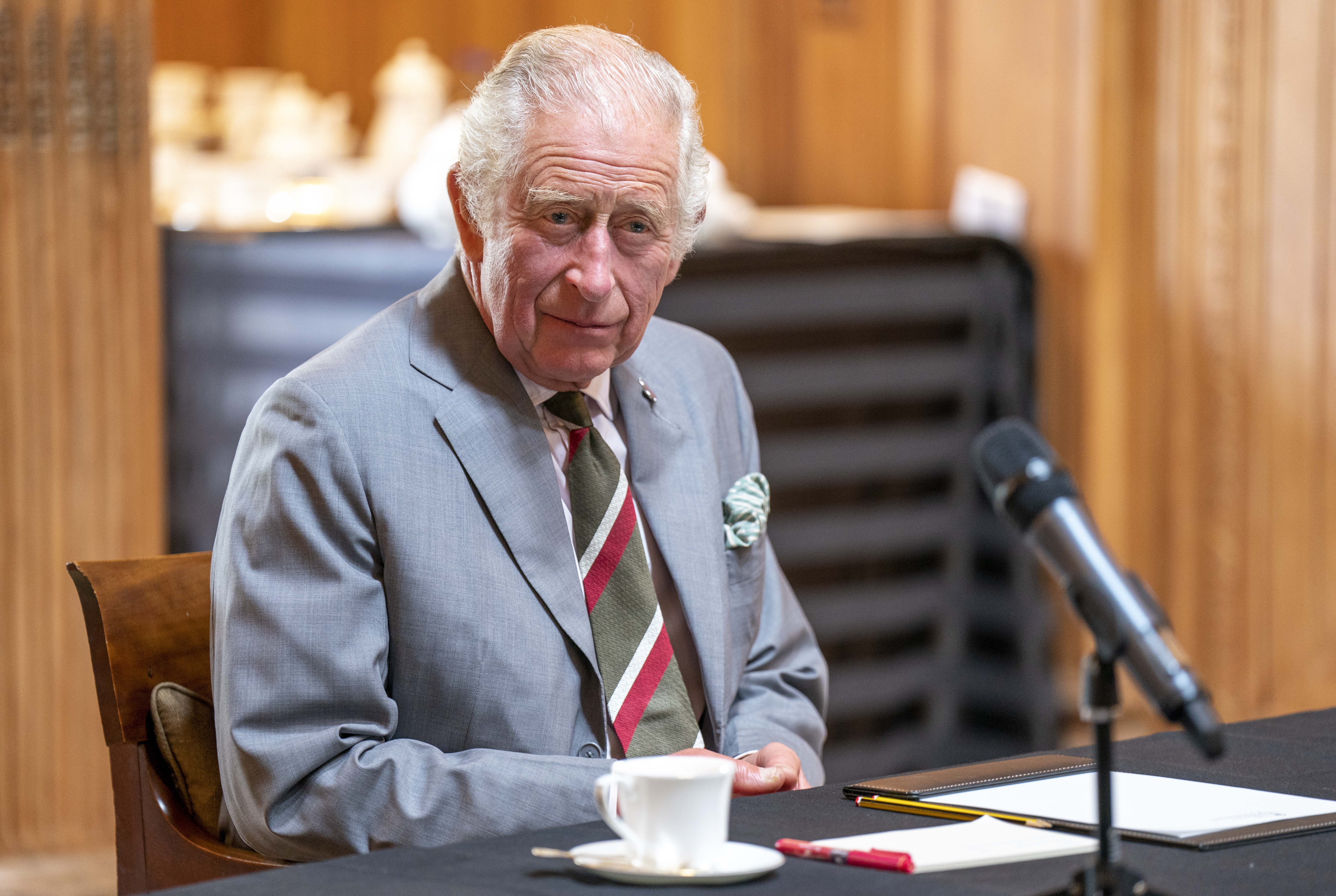 Prince Charles, in a gray suit and green-and-red striped tie, sits at a table in front of a microphone.