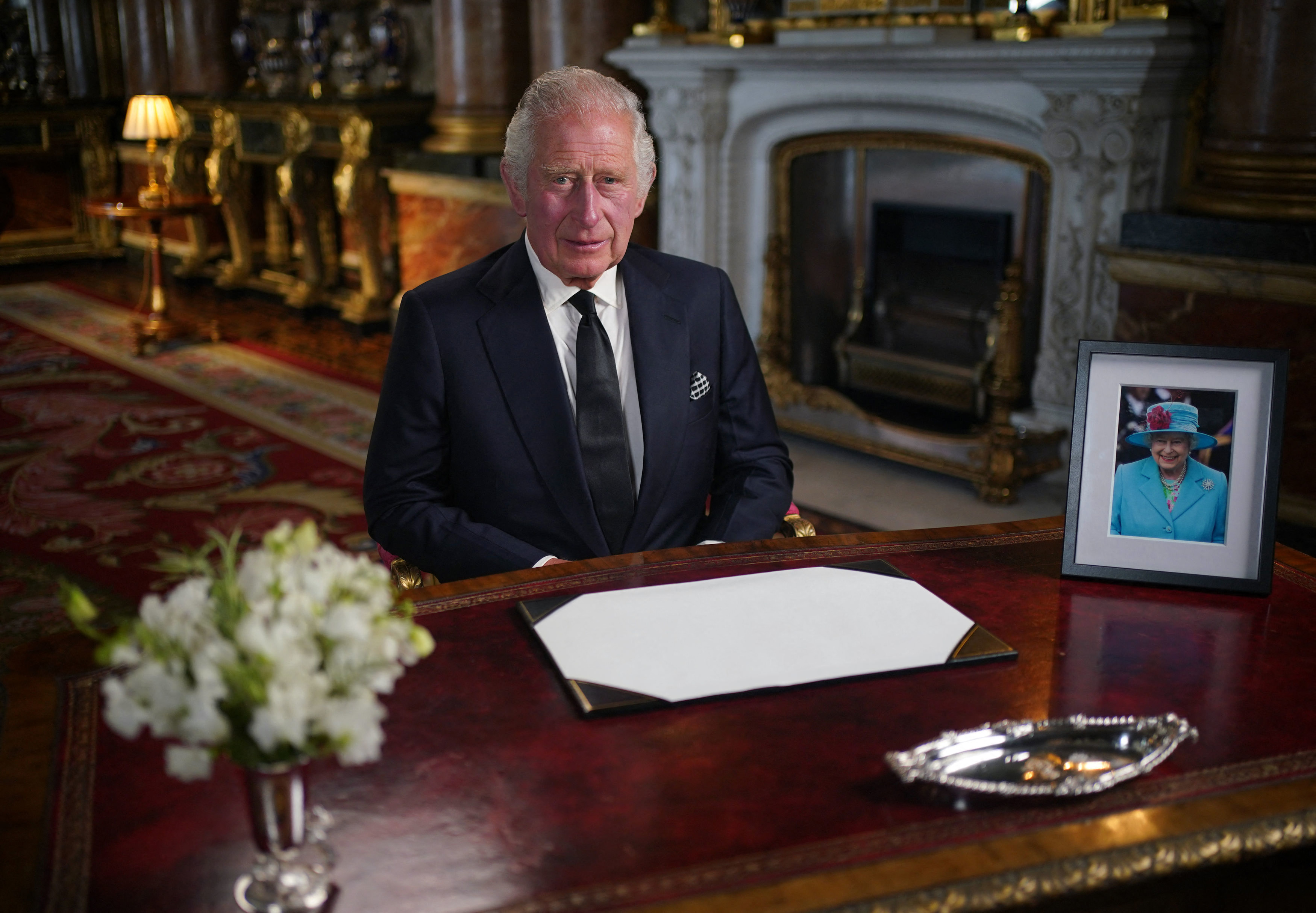 King Charles sits behind a desk with flowers and a picture of the late queen on it as he makes his televised speech.
