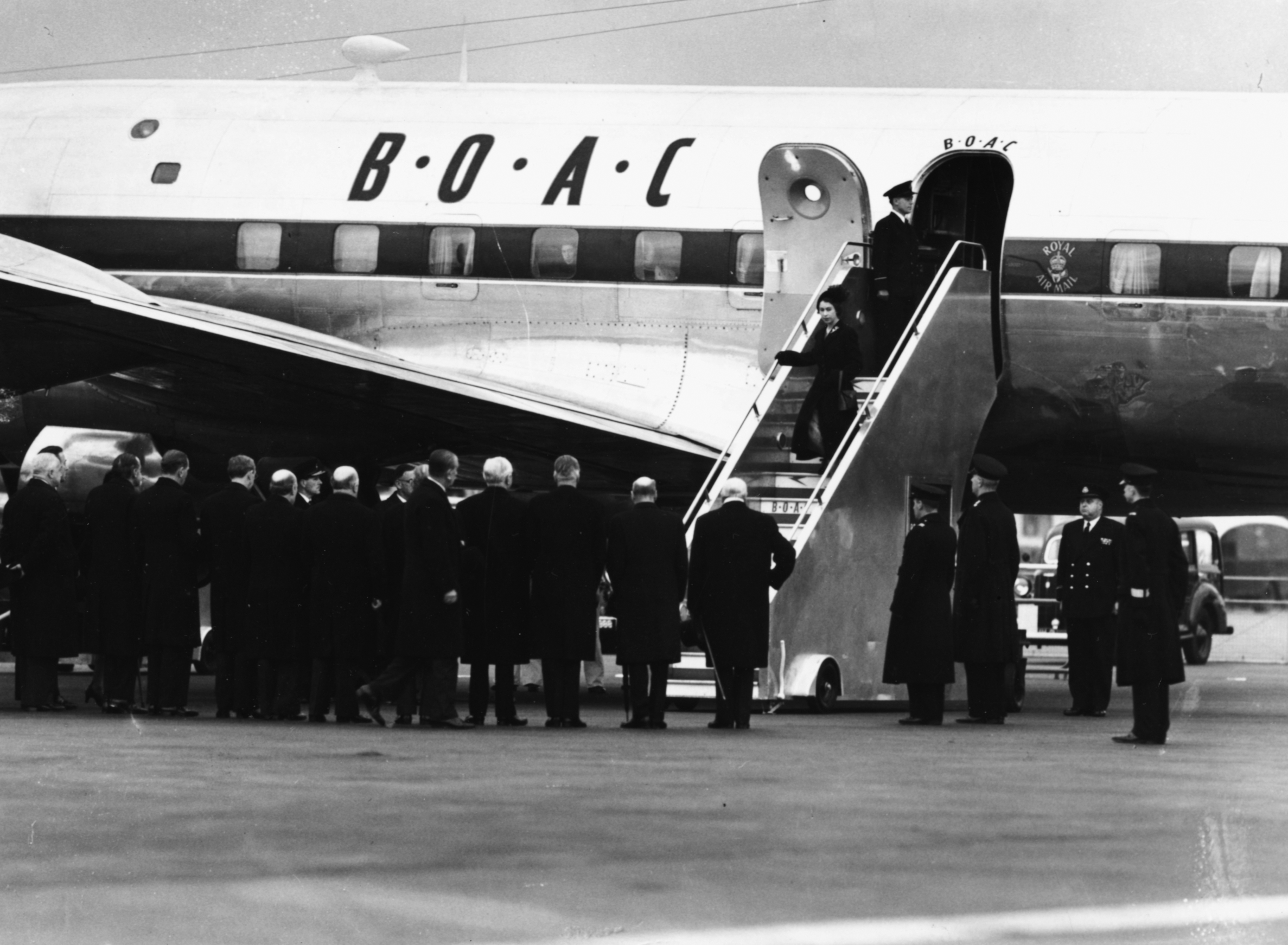 A black and white photo shows a man and woman walking down steps from an airplane as a small crowd waits. 