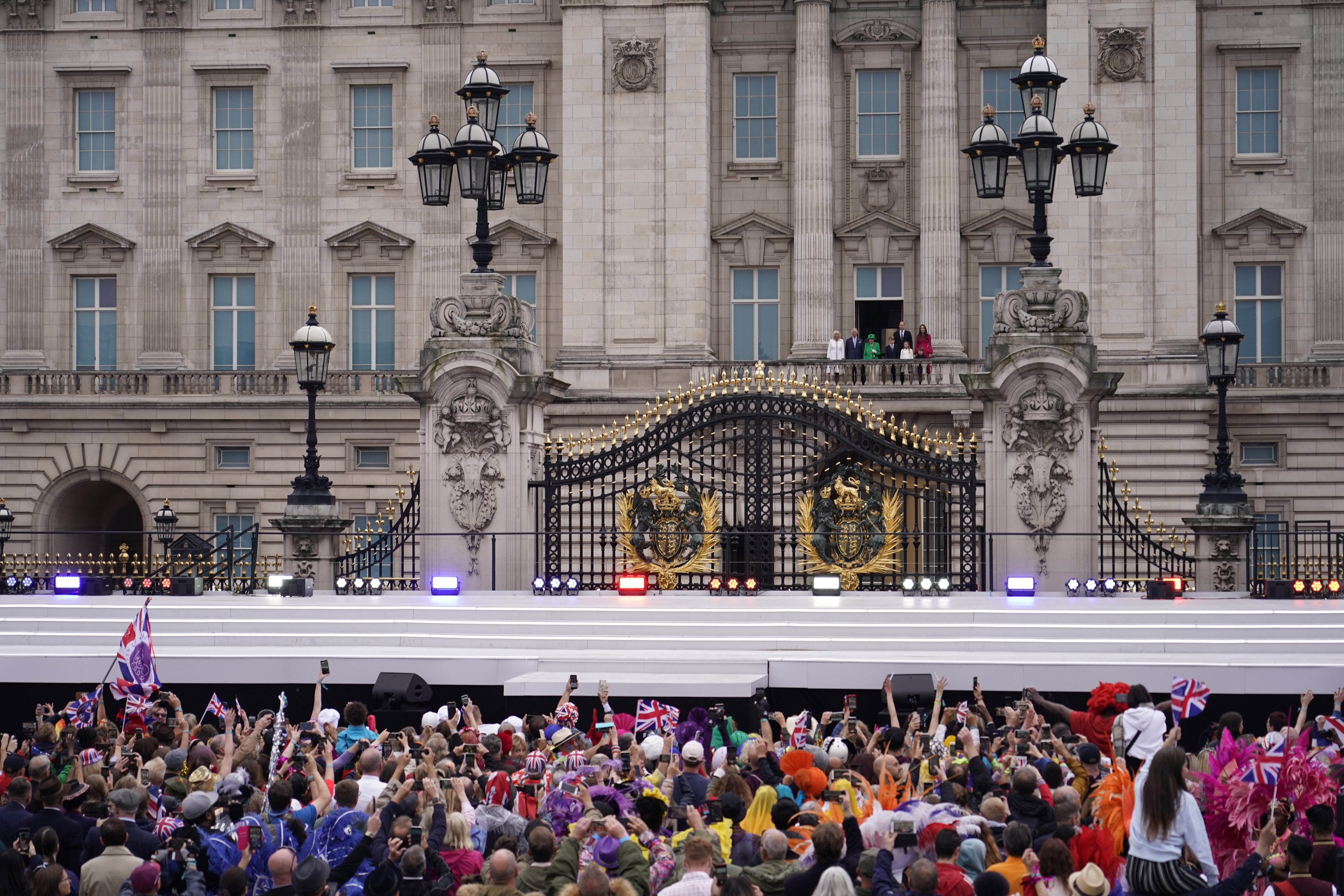 The Royal Family stands on a balcony of Buckingham Palace overlooking a large crowd of people waving flags. 