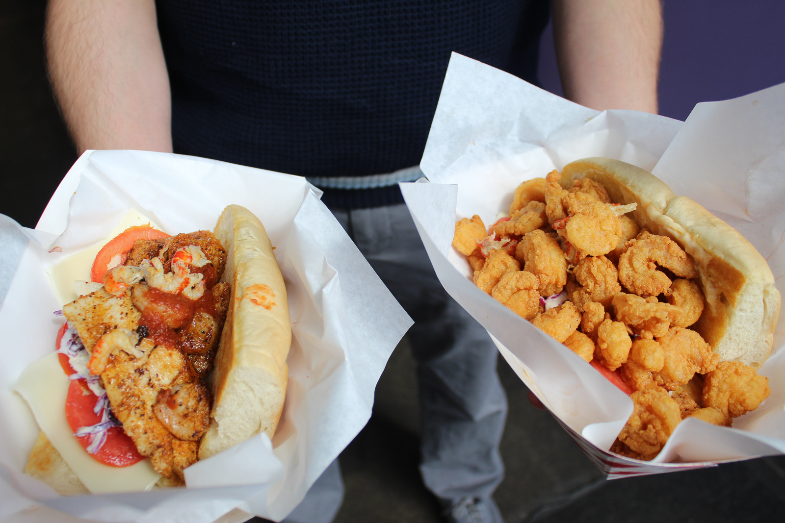 A person hold out two New Orleans-style po'boy sandwiches, one with grilled fish and the other with fried shrimp.