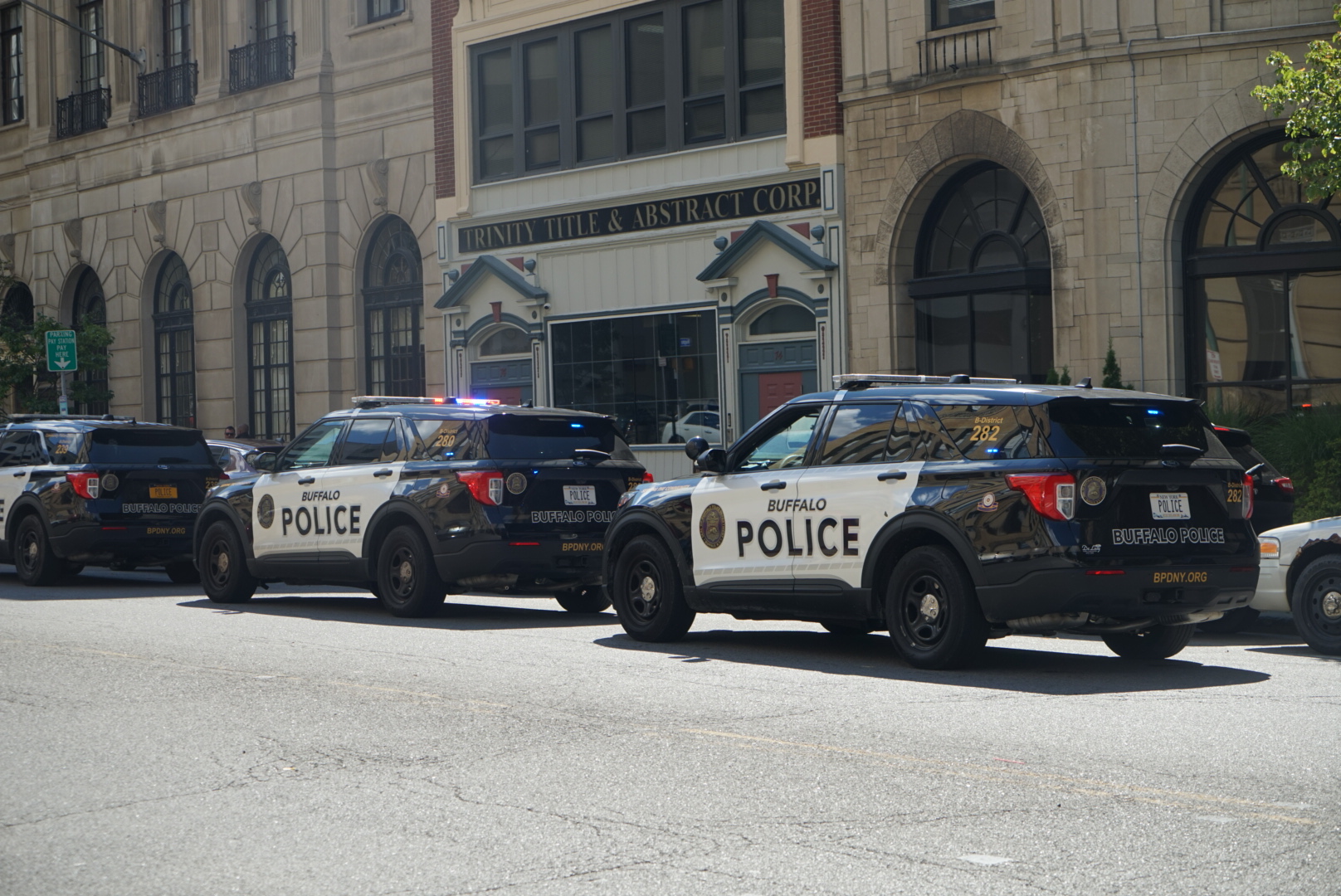 Three white and dark navy trucks lined along the road. The vehicles read "Buffalo Police". A building in the background reads "Trinity Title & Abstract Corp."