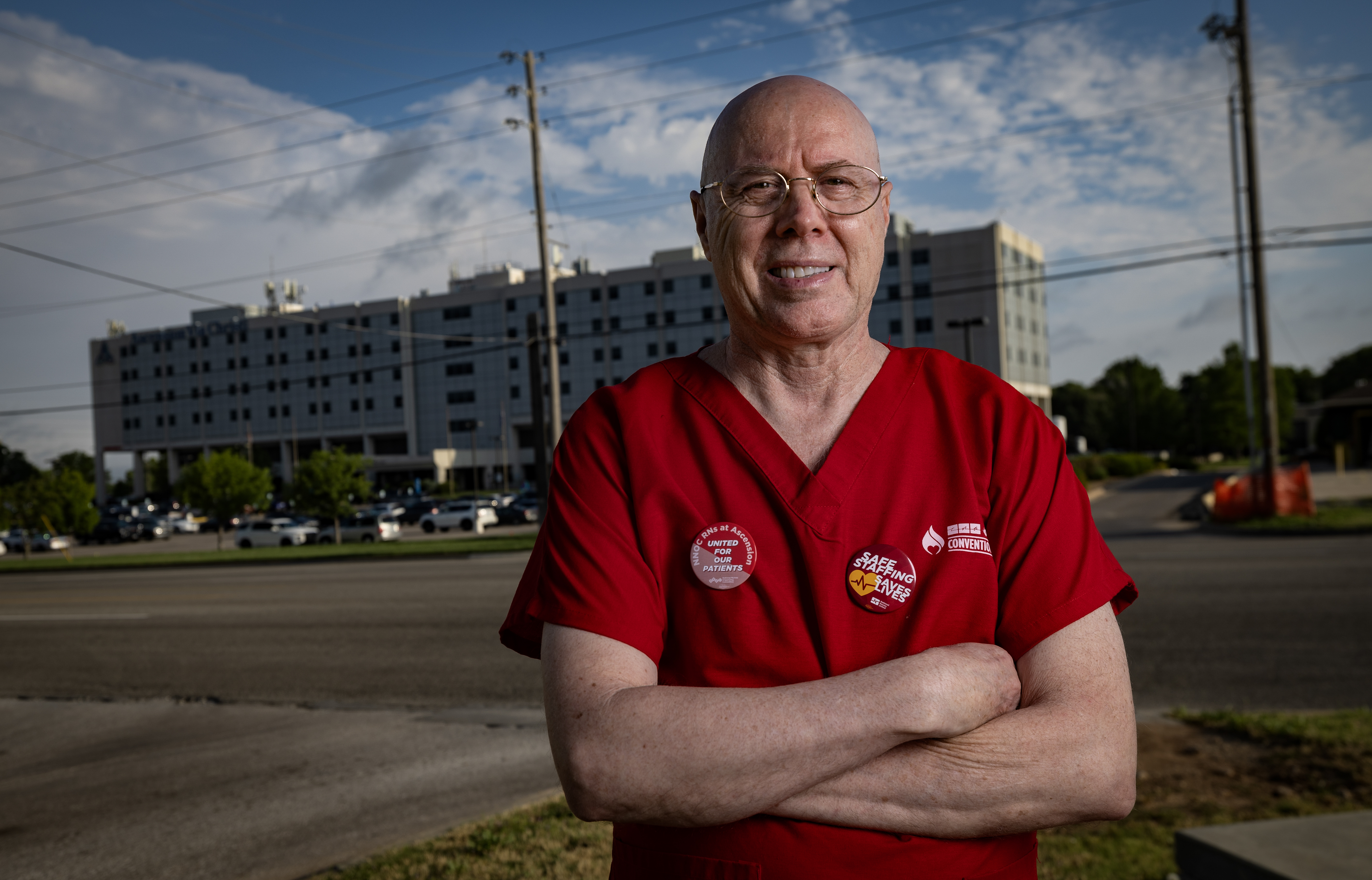 Marvin Ruckle, a middle aged man wearing glasses and red medical scrubs, stands in front of a hospital building with his arms folded in front of him. 