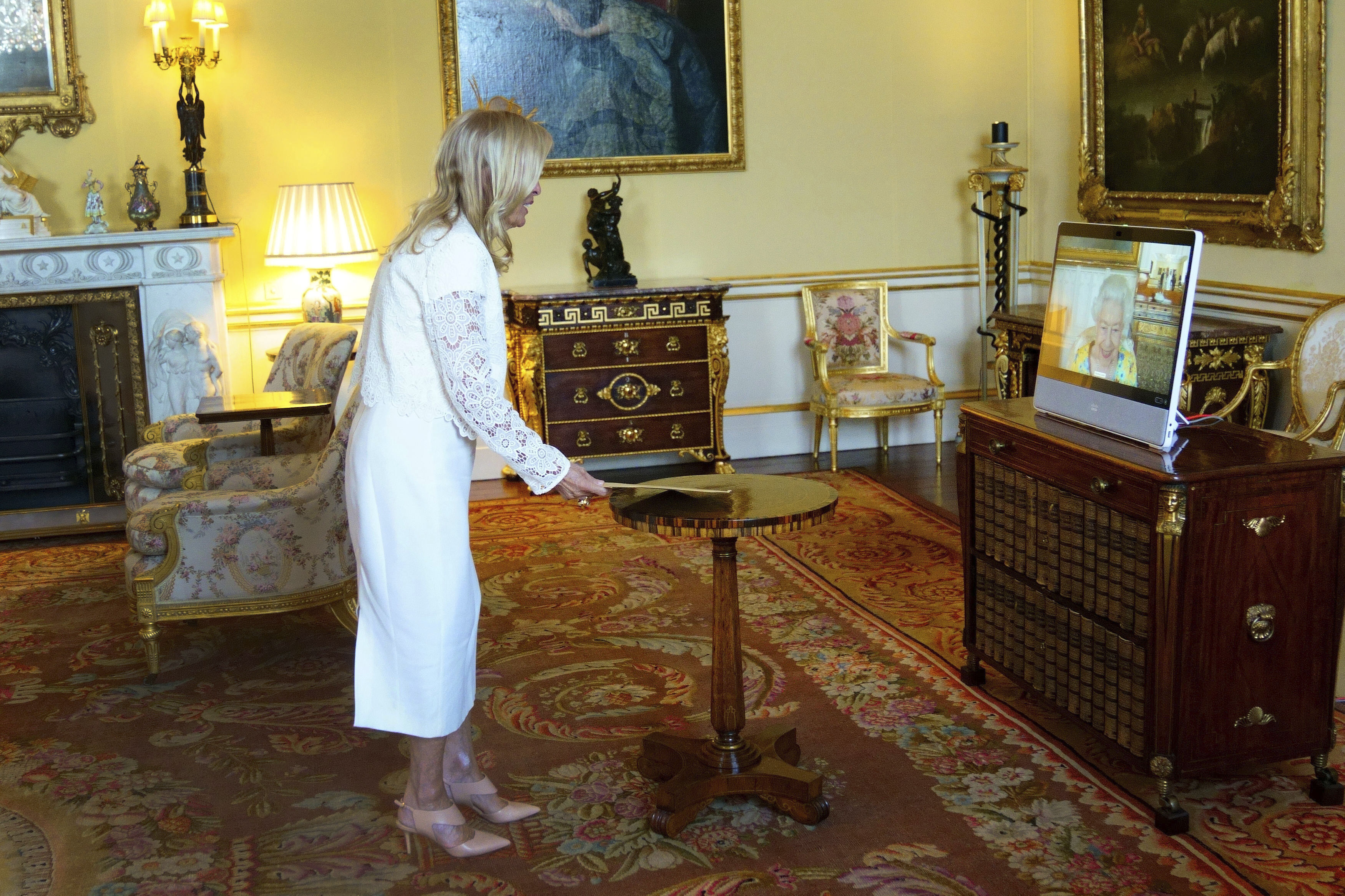 A blonde woman in a long-sleeve white dress stands in an ornate yellow and gold room in front of a computer screen showing a video call with Queen Elizabeth.