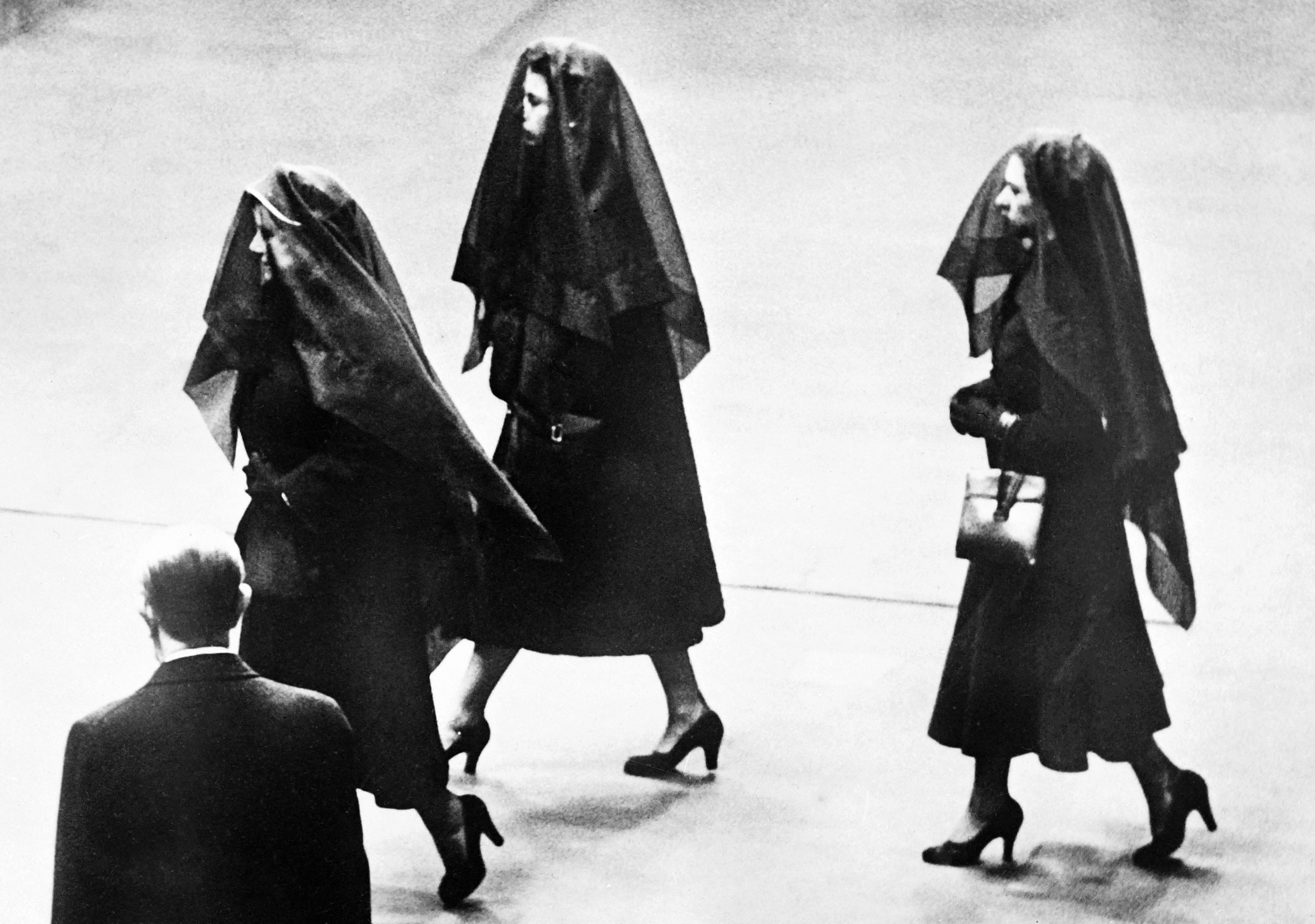 A black and white photo shows three women in dresses, heels and head coverings walking next to each other.