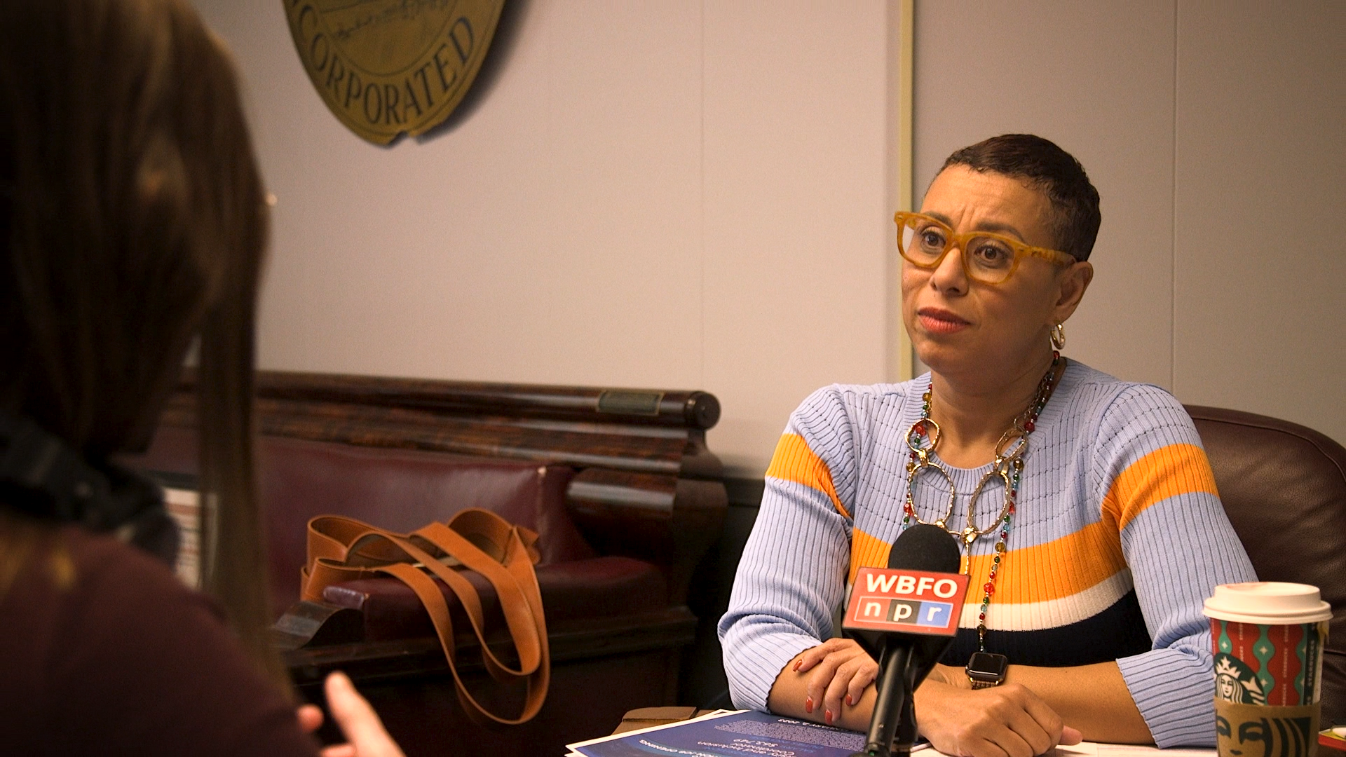 City of Buffalo Deputy Mayor Crystal Rodriguez-Dabney sits across from WBFO Disabilities Desk Reporter Emyle Watkins in her office. Rodriguez-Dabney is wearing brown glasses, earrings, necklaces and a blue, orange, white and black striped sweater. There is a WBFO microphone, papers, and a Starbucks cup in front of her.