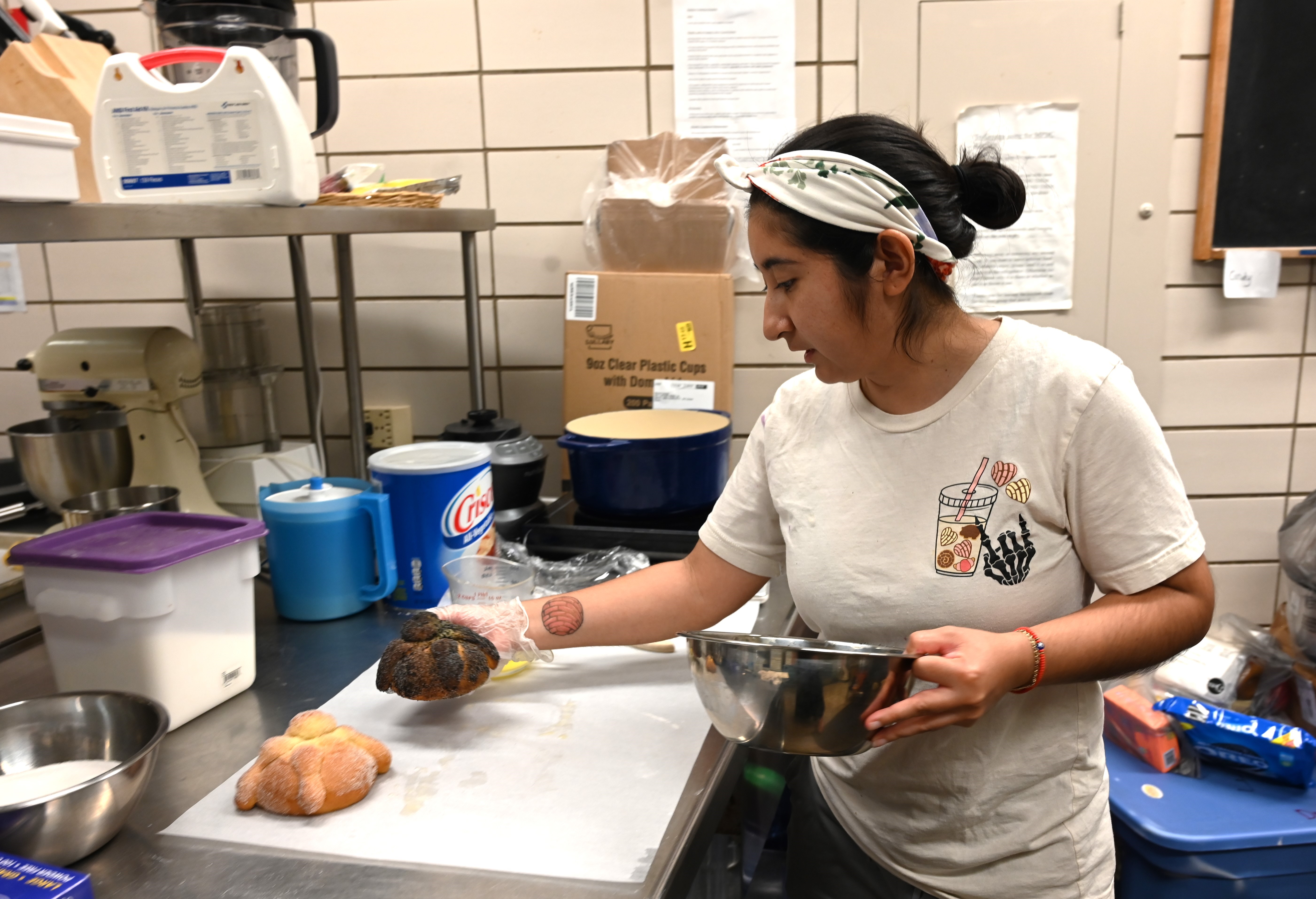 Norma places a fresh-baked pan de muerto, covered in corn husk ash and sugar, next to a traditional bread with sugar.