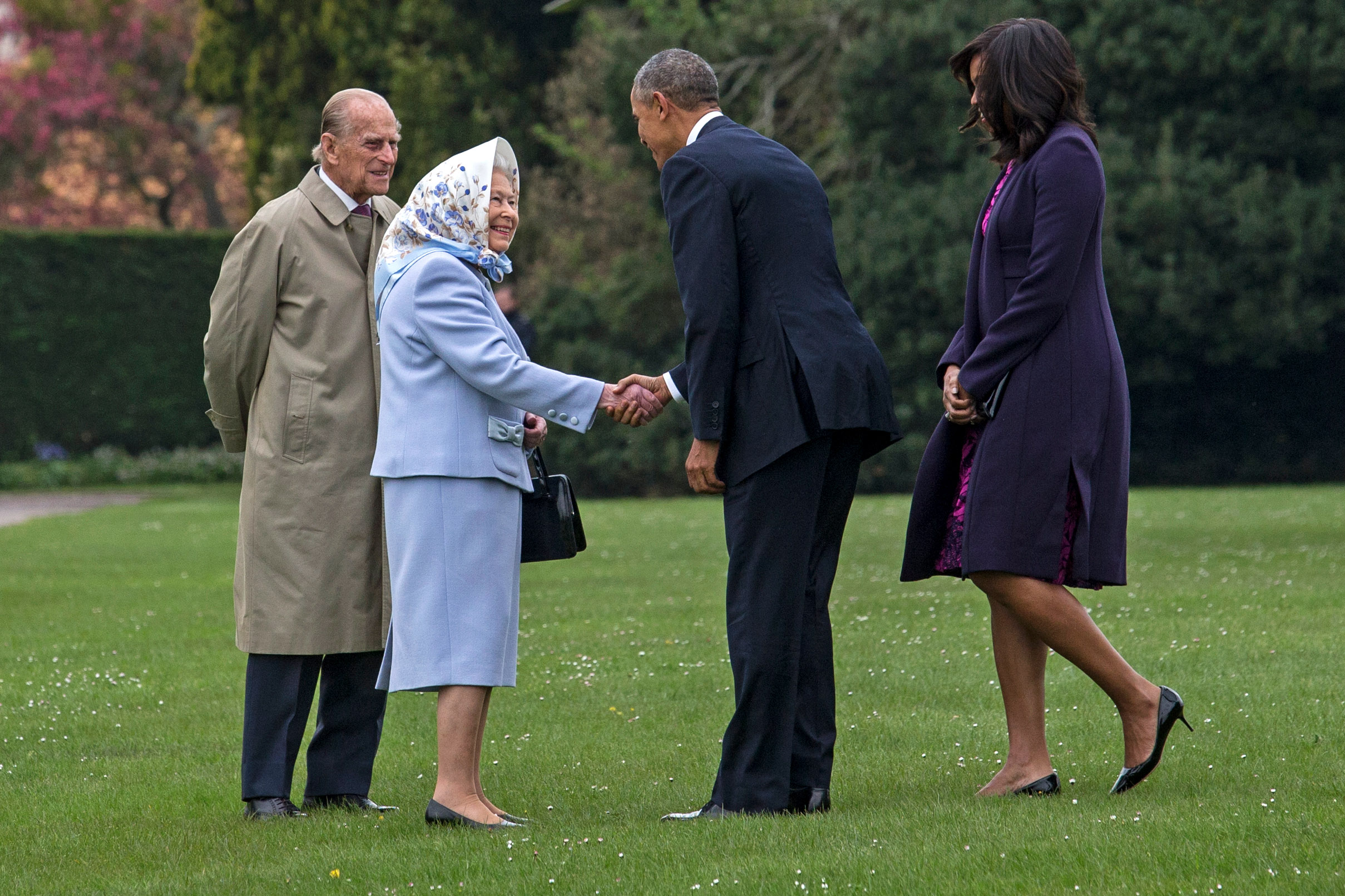 Queen Elizabeth, wearing a blue suit and headscarf, shakes hands with then-President Barack Obama as their spouses stand on the grass nearby.