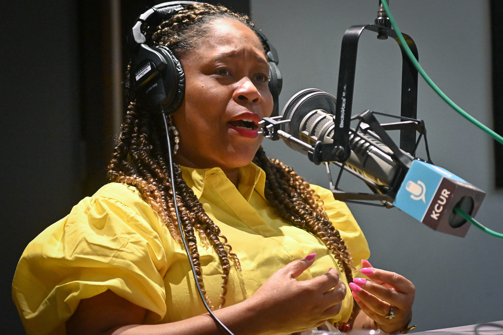 A woman wearing a bright yellow dress gestures with both hands while talking inside a studio at a microphone.